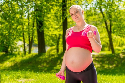 Active pregnant expectant mother with a dumbbell on a green lawn in a park