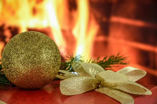 Red gift with a beautiful gold ribbon and a bauble on the background of a roaring fireplace
