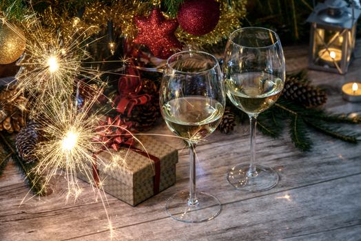 Sparklers at two wineglas with white wine on a wooden table in the background of Christmas tree, gift and candles