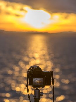 Cropped image with a modern DSLR camera, standing on a tripod, taking a photo with the sunset over the water, in live view mode.