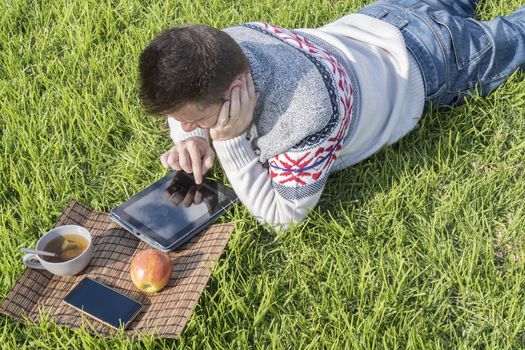 Man having breakfast lying on the grass in the garden with technology