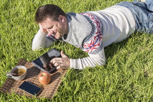 Man having breakfast lying on the grass in the garden with technology