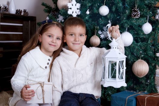 Two smiling kids sitting with lanterns under Christmas tree