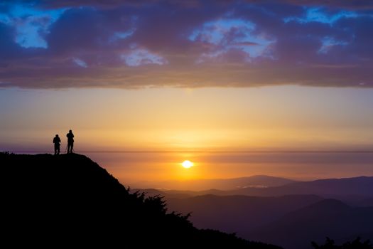 Silhouettes of two people standing on a rock and looking toward the sun. Dramatic clouds over them. Sunset in the mountains. Purple light. Summer in the Ukrainian Carpathians