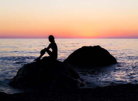 Silhouette of a girl that sitting on a rock by the sea and watching the sunset. Clear sky. Rest time by the sea in Georgia