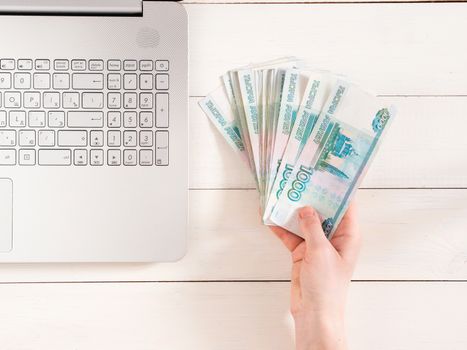 Hand holding russian rubles near laptop on white wooden background. Top view or flat lay. Finance and earning concept