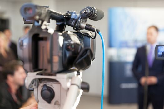 Video camera in focus, blurred spokesman in background. Corporate news conference.