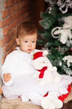 Portrait of a beautiful baby girl with a soft teddy bear in Santa hat in the interior with Christmas decorations.