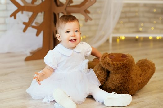 Portrait of a beautiful baby girl with a soft brown teddy bear in the interior with Christmas decorations.