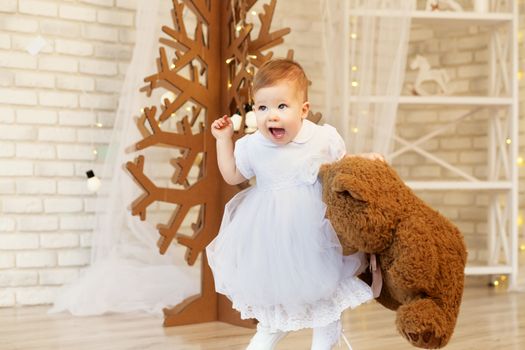 Portrait of a beautiful baby girl with a soft brown teddy bear in the interior with Christmas decorations.