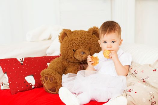 Child eats cookies. Portrait of a beautiful baby girl with a soft brown teddy bear in the interior with Christmas decorations.