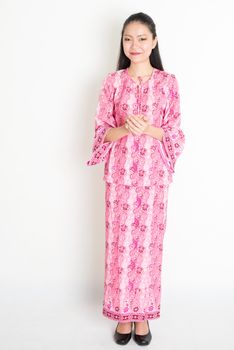 Portrait of young southeast Asian girl in traditional Malay batik dress greeting, standing on plain background.