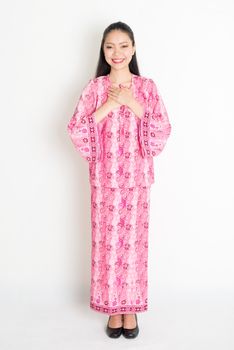 Portrait of young southeast Asian woman in traditional Malay batik dress greeting, standing on plain background.