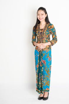Portrait of young southeast Asian woman in traditional Malay batik kebaya dress smiling, full length standing on plain background.