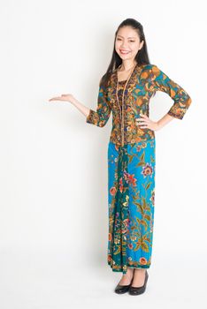 Portrait of young southeast Asian woman in traditional Malay batik kebaya dress smiling, full length standing on plain background.