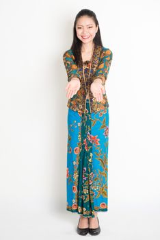 Portrait of young southeast Asian woman in traditional Malay batik kebaya dress hand holding something, full length standing on plain background.