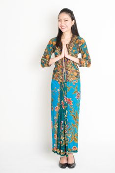 Portrait of young southeast Asian woman in traditional Malay batik kebaya dress greeting, full length standing on plain background.