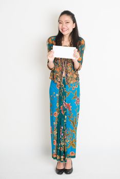 Portrait of young southeast Asian woman in traditional Malay batik kebaya dress hand holding a white blank paper card, full length standing on plain background.