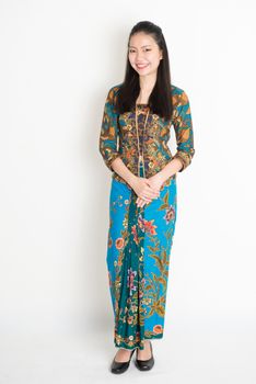 Portrait of young southeast Asian girl in traditional Malay batik kebaya dress smiling, full length standing on plain background.