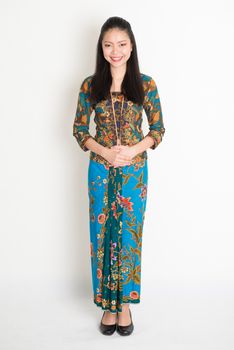 Portrait of young southeast Asian girl in traditional Malay batik kebaya dress smiling, full body standing on plain background.