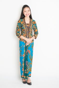 Portrait of young southeast Asian woman in traditional Malay batik kebaya dress smiling, full body standing on plain background.