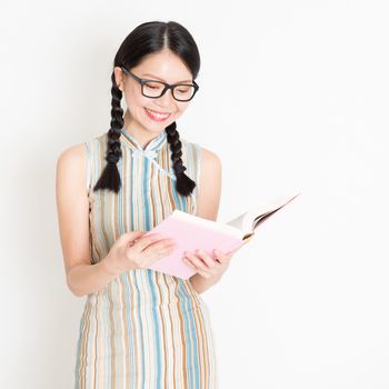 Portrait of young Asian girl in traditional qipao dress smiling and reading book, standing on plain background.