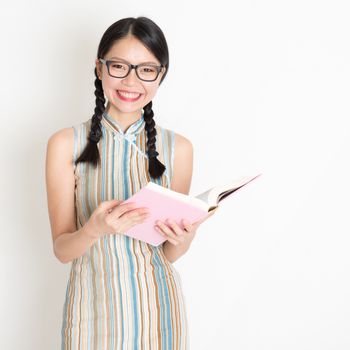 Portrait of young Asian woman in traditional qipao dress smiling and reading book, standing on plain background.