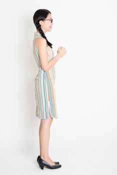 Side view or profile of young Asian girl in traditional qipao dress greeting, celebrating Chinese Lunar New Year, full length standing on plain background.