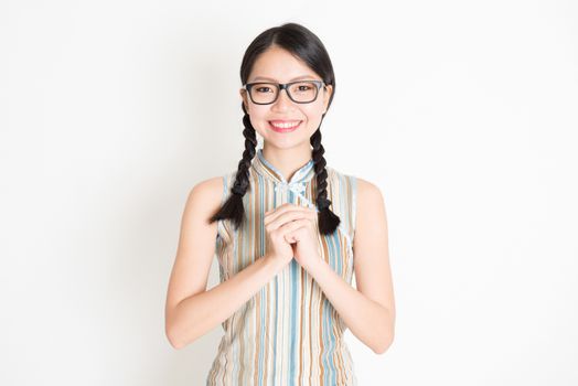 Portrait of young Asian girl in traditional qipao dress greeting, celebrating Chinese Lunar New Year or spring festival, standing on plain background.