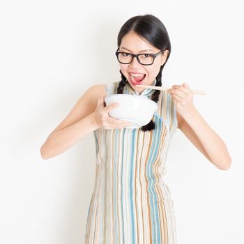 Portrait of young Asian girl in traditional qipao dress eating with chopsticks, celebrating Chinese Lunar New Year or spring festival, standing on plain background.
