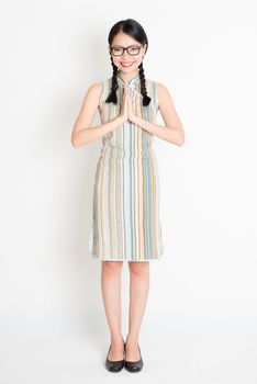 Portrait of young Asian girl in traditional qipao dress in greeting pose, full length standing on plain background.