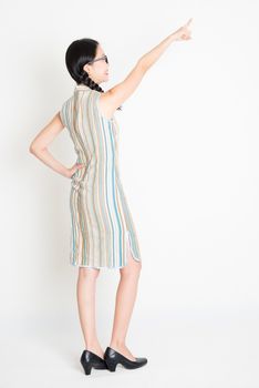 Rear view of young Asian girl in traditional qipao dress finger pointing away, full length standing on plain background.