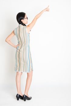 Rear view of young Asian woman in traditional qipao dress hand pointing away, full length standing on plain background.