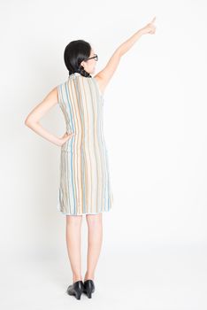 Rear view of young Asian girl in traditional qipao dress hand pointing away, full length standing on plain background.