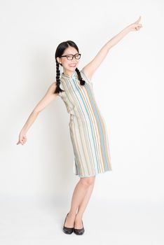 Portrait of young Asian girl in traditional qipao dress finger pointing at something, full length standing on plain background.