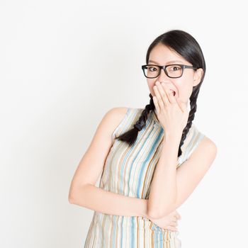 Portrait of young Asian girl in traditional qipao dress embarrassed and covering her mouth , standing on plain background.