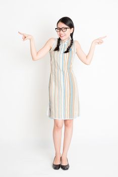 Portrait of young Asian woman in traditional qipao dress finger pointing at something, full length standing on plain background.