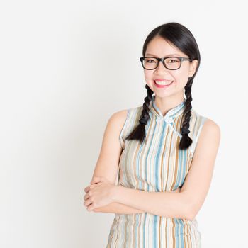 Portrait of young Asian female in traditional qipao dress smiling, celebrating Chinese Lunar New Year or spring festival, standing on plain background.