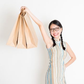 Portrait of young Asian female in traditional qipao dress shopping, hand holding paper bag, celebrating Chinese Lunar New Year or spring festival, standing on plain background.