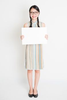 Portrait of young Asian woman in traditional qipao dress hand holding a white blank paper card, celebrating Chinese Lunar New Year or spring festival, full body standing on plain background.