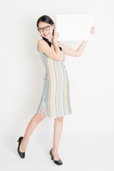 Portrait of young Asian girl in traditional qipao dress hand holding a white blank paper card, celebrating Chinese Lunar New Year or spring festival, full body standing on plain background.