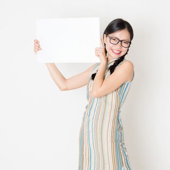 Portrait of young Asian girl in traditional qipao dress hand holding white blank paper card, celebrating Chinese Lunar New Year or spring festival, standing on plain background.