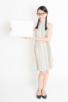Portrait of young Asian woman in traditional qipao dress hand holding white blank paper card, celebrating Chinese Lunar New Year or spring festival, full body standing on plain background.