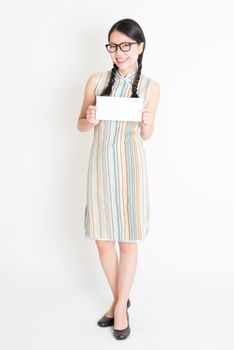 Portrait of young Asian female in traditional qipao dress hand holding white blank paper card, celebrating Chinese Lunar New Year or spring festival, full body standing on plain background.