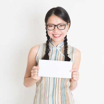Portrait of young Asian girl in traditional cheongsam dress hand holding white blank paper card, celebrating Chinese Lunar New Year or spring festival, standing on plain background.