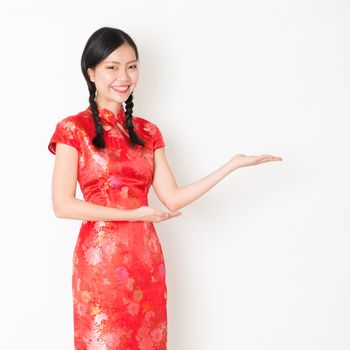 Portrait of young Asian woman in traditional qipao dress smiling and hand showing something, celebrating Chinese Lunar New Year or spring festival, standing on plain background.