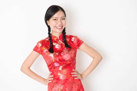 Portrait of young Asian girl in traditional qipao dress smiling, celebrating Chinese Lunar New Year or spring festival, standing on plain background.