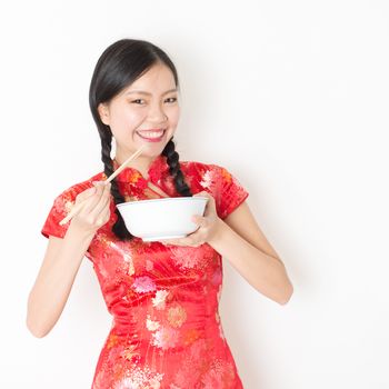 Portrait of young Asian woman in traditional qipao dress eating, hand holding bowl and chopsticks, celebrating Chinese Lunar New Year or spring festival, standing on plain background.