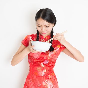 Portrait of young Asian female in traditional qipao dress eating, hand holding bowl and chopsticks, celebrating Chinese Lunar New Year or spring festival, standing on plain background.