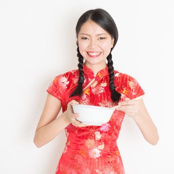 Portrait of young Asian girl in traditional qipao dress eating, hand holding bowl and chopsticks, celebrating Chinese Lunar New Year or spring festival, standing on plain background.